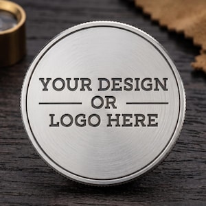 May include: A circular, brushed silver metal coin or token featuring a textured, knurled edge. The center is engraved with the text "YOUR DESIGN OR LOGO HERE" in a bold, sans-serif font, arranged in three lines with horizontal lines separating them.