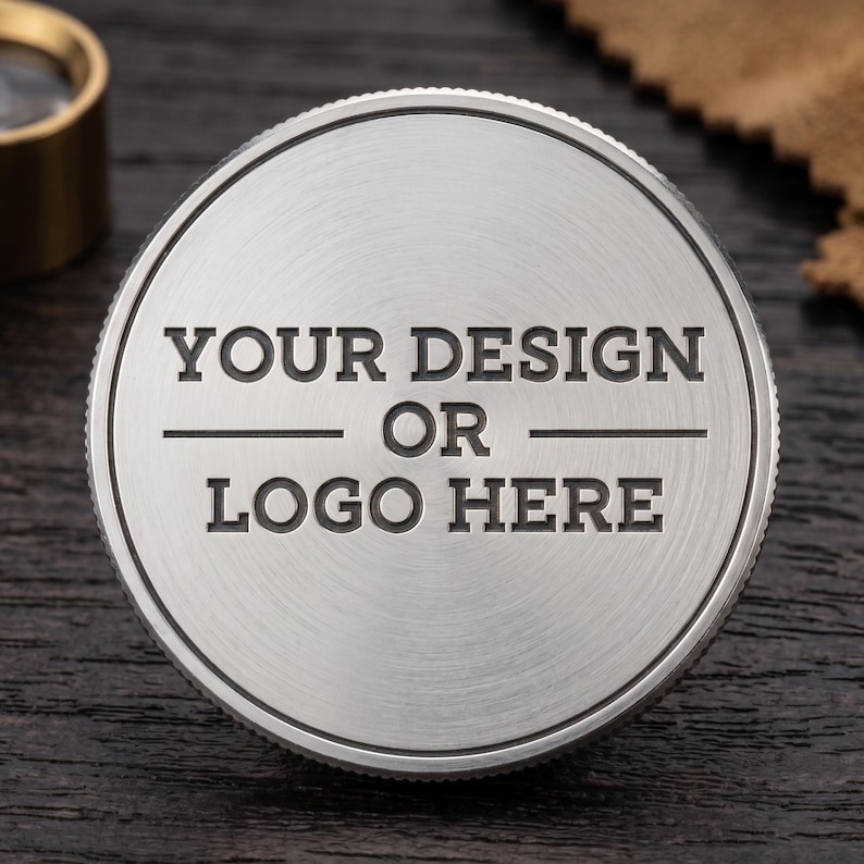 May include: A circular, brushed silver metal coin or token featuring a textured, knurled edge. The center is engraved with the text "YOUR DESIGN OR LOGO HERE" in a bold, sans-serif font, arranged in three lines with horizontal lines separating them.