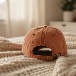 May include: A terracotta baseball cap, seen from behind, with a curved peak and metal clasp. The cap sits on a cream-coloured, textured blanket. The stitching and construction of the cap are clearly visible. The cap is a warm, earthy tone.