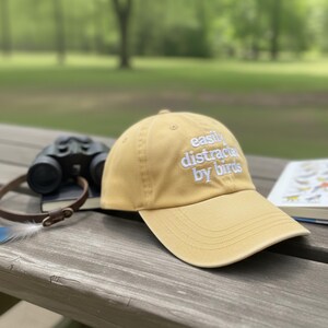 May include: A mustard-colored baseball cap with the embroidered text "easily distracted by birds." The hat is angled on a wooden picnic table, with binoculars and a bird book in the background. The scene is set outdoors with a blurred green backdrop.