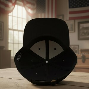 May include: A black baseball cap with a curved brim, viewed from the underside. The interior is light gray with a white center button. The cap has a brown adjustable strap. The background includes American flags and a wooden surface.