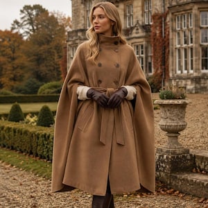 May include: A camel-colored double-breasted wool cape coat featuring a high collar, a matching tie belt at the waist, and side pockets. The garment is worn with dark brown leather gloves, a cream-colored knit sweater, and dark brown leather boots.