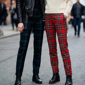 May include: Two people wearing stylish outfits. One wears a black leather jacket and green plaid pants. The other wears a cream-colored sweater and red plaid pants. Both wear black boots. The setting appears to be an urban street.