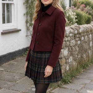 May include: A burgundy wool-blend jacket with a collar and three front buttons, paired with a dark green and navy blue plaid pleated skirt, a black turtleneck sweater, black opaque tights, and black knee-high boots.