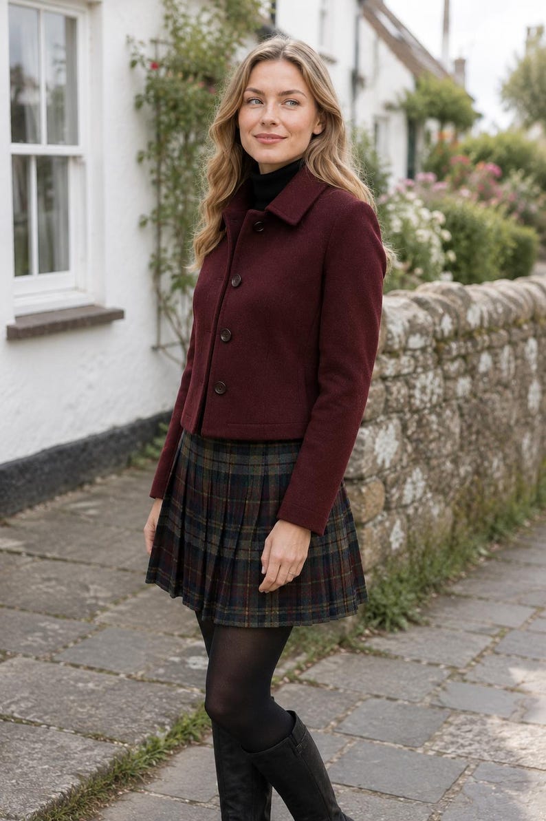 May include: A burgundy wool-blend jacket with a collar and three front buttons, paired with a dark green and navy blue plaid pleated skirt, a black turtleneck sweater, black opaque tights, and black knee-high boots.