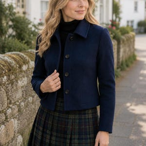 May include: A navy blue cropped jacket with a collar and four dark buttons down the front, worn over a black turtleneck sweater and a dark green and navy blue plaid pleated skirt with thin red lines throughout the pattern.