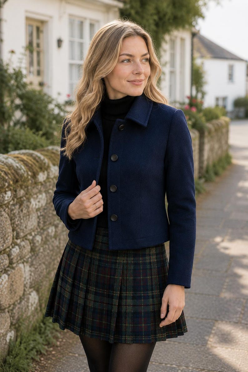 May include: A navy blue cropped jacket with a collar and four dark buttons down the front, worn over a black turtleneck sweater and a dark green and navy blue plaid pleated skirt with thin red lines throughout the pattern.