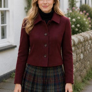 May include: A burgundy button-up jacket with a collar and long sleeves, worn over a black turtleneck sweater and a pleated plaid skirt featuring green, blue, and red tones. The jacket has three visible buttons down the front center.