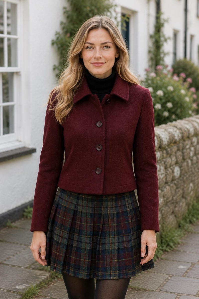 May include: A burgundy button-up jacket with a collar and long sleeves, worn over a black turtleneck sweater and a pleated plaid skirt featuring green, blue, and red tones. The jacket has three visible buttons down the front center.
