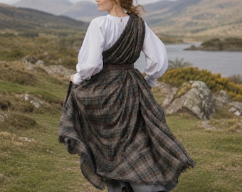 May include: A woman wearing a white blouse with long sleeves and a plaid skirt. The skirt is gray, brown, and red. A brown sash is around the waist. The woman is running on a grassy hill with a lake and mountains in the background.