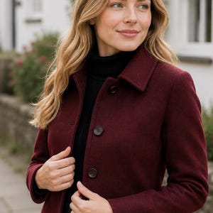 May include: A deep burgundy, hip-length jacket featuring a structured collar, long sleeves, and a front button closure with three dark buttons. The jacket is worn over a black ribbed turtleneck sweater and a dark green and navy plaid pleated skirt.