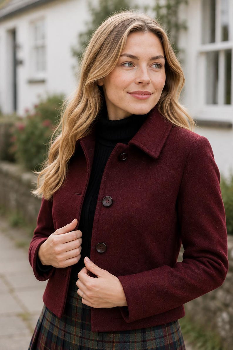 May include: A deep burgundy, hip-length jacket featuring a structured collar, long sleeves, and a front button closure with three dark buttons. The jacket is worn over a black ribbed turtleneck sweater and a dark green and navy plaid pleated skirt.