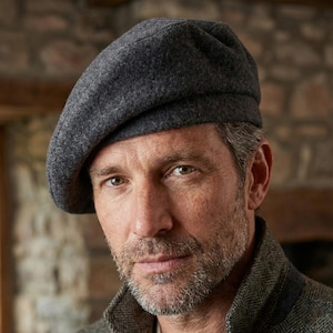 May include: A dark gray beret made of a soft, textured material. The hat is worn at an angle, with the brim resting on the forehead. The beret is a classic style, suitable for various occasions.