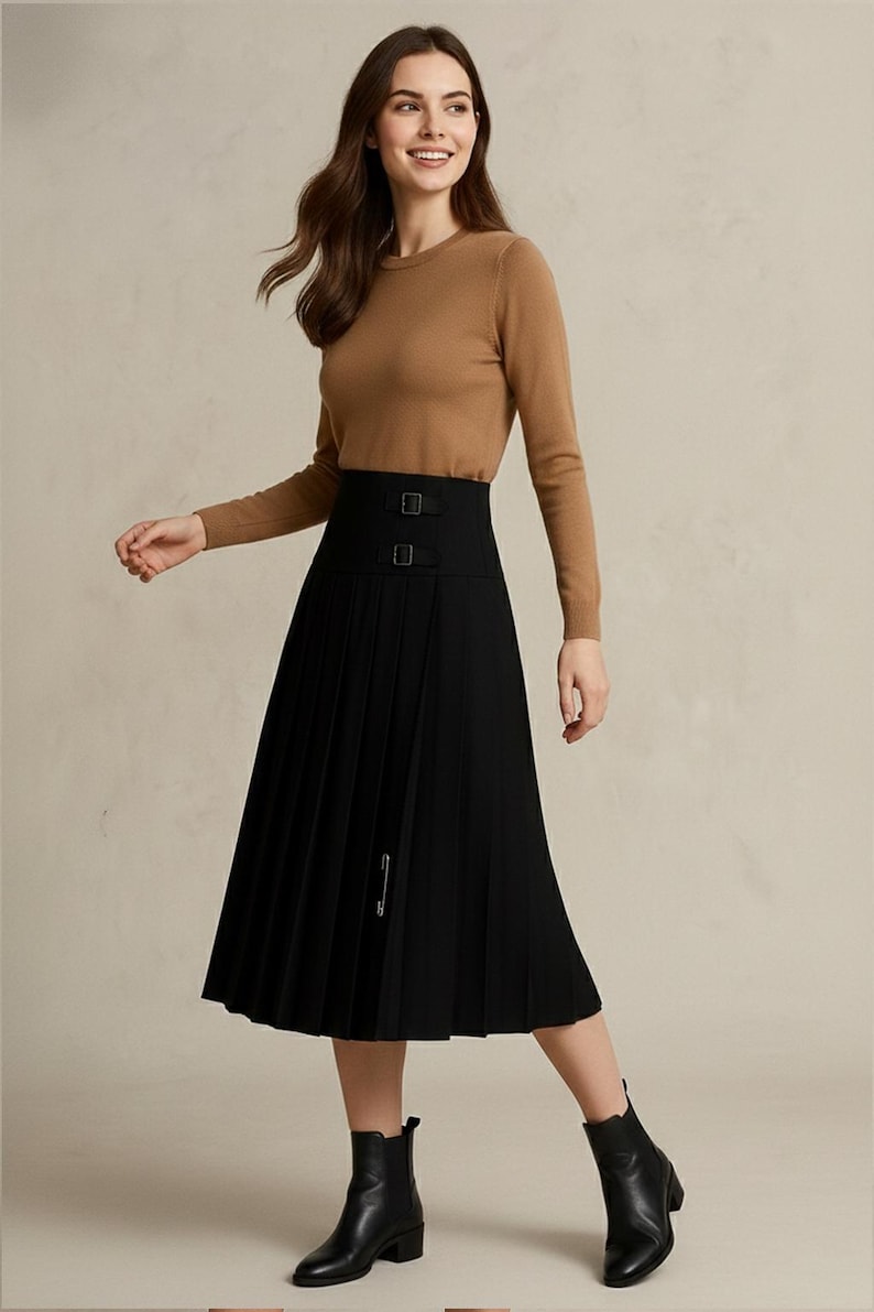 May include: A woman wearing a brown long-sleeve sweater and a black pleated skirt with a high waist and buckle details. The skirt falls to mid-calf length. She is wearing black ankle boots.