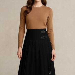 May include: A woman wearing a brown long-sleeve sweater and a black pleated skirt. The skirt has a high waist with two buckle details and a safety pin. The outfit is a classic and elegant style.