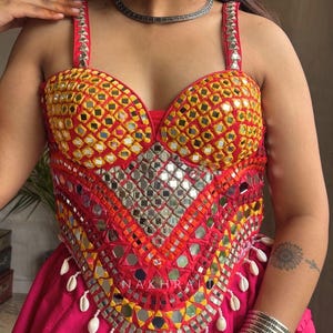May include: A sleeveless corset top in vibrant red and yellow, heavily embellished with small square mirrors and intricate embroidery. The hem features dangling white cowrie shells, and the word NAKHRA is printed in white on the front panel.