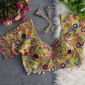 May include: A vibrant yellow blouse featuring intricate multi-colored geometric embroidery, circular mirror accents, and a deep neckline with tie-up strings. The hem is adorned with small white hanging shells, creating a detailed and textured garment.