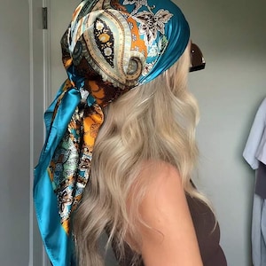 Puede incluir: Un pañuelo de seda turquesa con un estampado de cachemira, atado alrededor de la cabeza. El pañuelo presenta una mezcla de colores turquesa, naranja y crema. El pañuelo está atado en la nuca, con los extremos colgando.