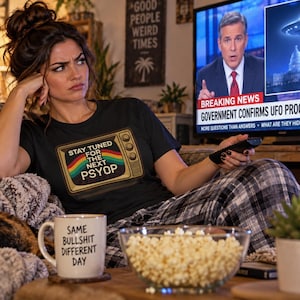 May include: A black t-shirt featuring a graphic of a vintage television set with the text "STAY TUNED FOR THE NEXT PSYOP" and a rainbow stripe. The shirt is paired with black and white plaid pajama pants and a white mug with "SAME BULLSHIT DIFFERENT DAY" printed on it.