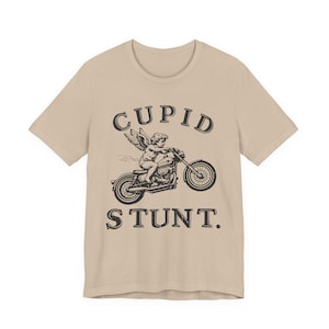 Op de afbeelding: Beige t-shirt met een zwarte afbeelding van Cupido op een motor. De woorden "CUPID STUNT" staan boven en onder de afbeelding. Het shirt heeft een ronde hals en korte mouwen.