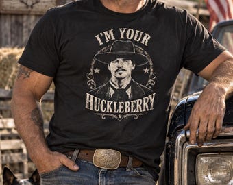 Può includere: Una t-shirt nera a maniche corte con una grafica bianca dall'aspetto vissuto raffigurante un uomo con un cappello da cowboy e la scritta I'M YOUR HUCKLEBERRY. Il design include un bordo circolare con stelle e decorazioni stampate sul tessuto.