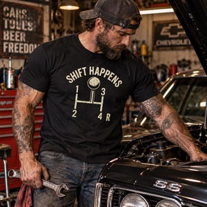 May include: A black short-sleeved t-shirt featuring a cream-coloured graphic of a manual transmission gear shift pattern with the text SHIFT HAPPENS above it. The design includes the numbers 1, 2, 3, 4 and the letter R arranged in a standard H-pattern.