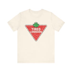 Puede incluir: Camiseta color crema con un triángulo rojo desgastado que lleva el texto "TIRED CANADIAN" y una hoja de arce verde. El gráfico tiene un aspecto vintage y desgastado, lo que sugiere un estilo informal.