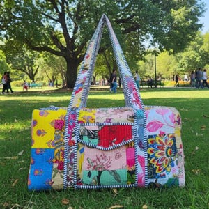 May include: A colorful patchwork tote bag with a rectangular body and triangular handles. The bag features a variety of floral and geometric patterns in shades of yellow, blue, pink, and red. The bag is sitting on green grass.
