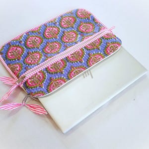 May include: A quilted laptop sleeve with a floral pattern in pink, green, and gold on a blue background. The sleeve has a pink zipper and trim, with pink and white striped ribbon zipper pulls. A white laptop is partially visible.