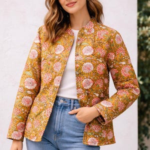 May include: Mustard-colored quilted jacket with a floral pattern in pink and white. The jacket has a mandarin collar and is worn over a white top and blue jeans. The jacket is a lightweight, casual outerwear piece.