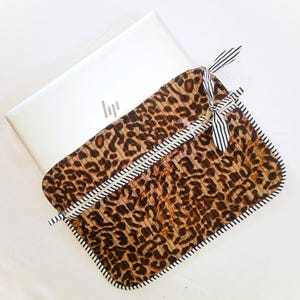 May include: A leopard print laptop sleeve with a black and white striped ribbon closure. The sleeve is brown with black spots and a white zipper. The laptop is white with the HP logo.