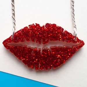 Puede incluir: Un collar de cadena plateada con un colgante de labios de acrílico rojo cubierto de purpurina.