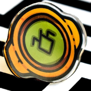May include: A circular pin with a green central design and orange and brown concentric circles. The pin has a black outline and sits against a black and white zigzag pattern. The central design is a stylized symbol.