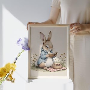 May include: A framed watercolor print featuring a bunny in a blue jacket reading a book. The bunny sits on a grassy area with a bowl of food and a red and white checkered blanket. The artwork has a soft, pastel color palette.