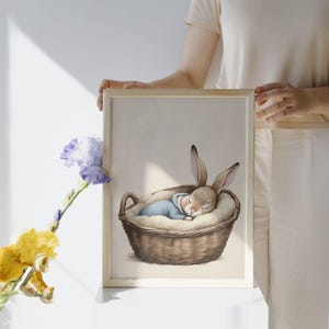 Könnte beinhalten: Ein gerahmter Aquarelldruck zeigt ein schlafendes Kaninchen in einem geflochtenen Korb. Das Kaninchen ist hellbraun mit langen Ohren und trägt einen blauen Anzug. Der Korb ist braun und mit weißem Flaum gefüttert.
