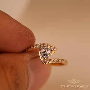 May include: A gold ring with a unique design, featuring a cluster of clear gemstones at the center and smaller stones along the band. The ring is held by a person's finger, showcasing its intricate details and craftsmanship. The ring is from Stamford Jewels.