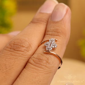 May include: A rose gold ring with a cluster of small, sparkling white stones forming a floral design. The ring is on a finger, showcasing the delicate details and elegant style. The ring is from Stamford Jewels.