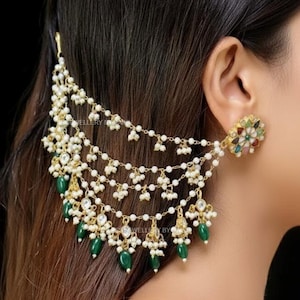 Pode incluir: Um conjunto de brincos e acessórios de cabelo dourados ornamentados. O brinco apresenta um design floral com pedras preciosas coloridas. O acessório de cabelo tem vários fios de pérolas e contas verdes em forma de gota, criando um efeito em cascata. O texto "Reference Photo" é visível.