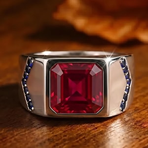 Puede incluir: Un anillo de tono plateado que presenta una gran piedra preciosa de color rojo intenso y corte cuadrado en el centro. La banda está acentuada a ambos lados por una fila vertical de pequeñas piedras preciosas redondas de color azul oscuro.