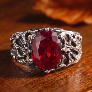 May include: A silver ring featuring a large, oval-cut, deep red gemstone set in a band with a textured, organic, branch-like metal design. The metal has a dark, oxidized finish that highlights the intricate, irregular patterns surrounding the stone.