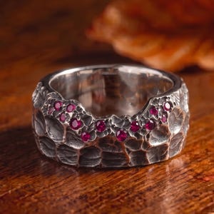 Może przedstawiać: Szeroki srebrny pierścionek o mocno teksturowanej, młotkowanej powierzchni z ciemnym, oksydowanym wykończeniem. Szereg małych, okrągłych, jaskrawoczerwonych kamieni szlachetnych jest osadzony wzdłuż górnej krawędzi obrączki w nieregularny, falisty wzór.