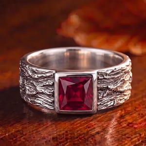 May include: A silver-toned ring featuring a square-cut, deep red gemstone set in the centre. The band of the ring has a textured, uneven surface that mimics the appearance of tree bark, contrasting with the smooth, polished metal setting.