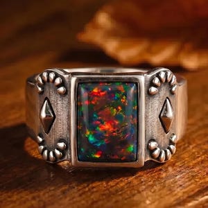 May include: A silver ring featuring a rectangular black opal with vibrant red, green, and blue flashes. The band has an oxidized finish with embossed decorative patterns, including small circular accents and a central diamond shape on each side.