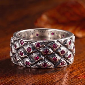 Op de afbeelding: Een brede zilveren ring met een getextureerd patroon van overlappende schubben op het buitenoppervlak. Elke schub is voorzien van een kleine, ronde, dieprode edelsteen in het midden, wat zorgt voor een gedetailleerd en herhalend ontwerp.