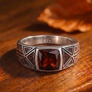 Könnte beinhalten: Ein silberner Ring mit einem großen, quadratischen, roten Edelstein in der Mitte. Der Ring hat ein geometrisches Design am Band und die Zahl "925" ist sichtbar. Der Ring wird auf einer Holzoberfläche präsentiert.