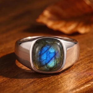 May include: A silver-toned metal ring featuring a smooth, polished band and a square-shaped, bezel-set labradorite gemstone. The stone displays a prominent iridescent blue flash across its surface, contrasting with the metallic luster of the band.