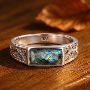 May include: A sterling silver ring featuring a rectangular labradorite gemstone with blue and gold iridescence. The band is engraved with intricate leaf patterns on both sides and is stamped with the number 925 on the inner surface.