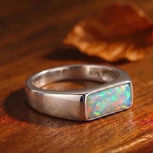 May include: A silver ring featuring a rectangular opal gemstone. The opal showcases a vibrant display of colour, including green, blue, and orange tones. The ring is set on a wooden surface, with a blurred leaf in the background.
