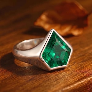 May include: A silver ring featuring a large, emerald-cut green gemstone. The ring has a wide, polished band and a bezel setting that highlights the vibrant green color of the stone. The ring is displayed on a wooden surface.