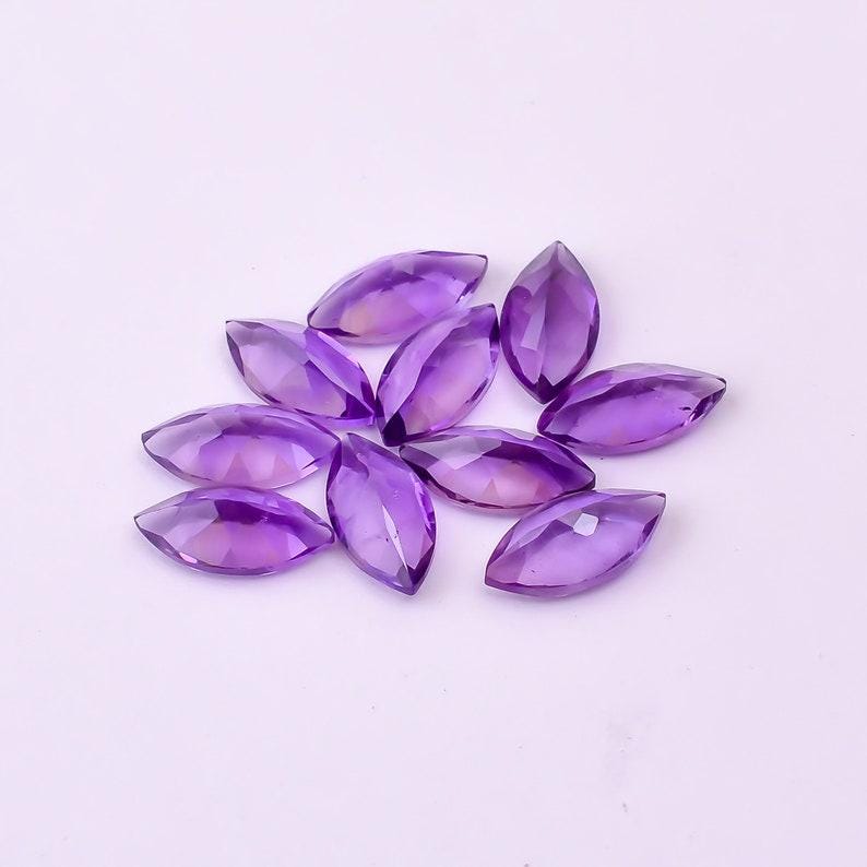 May include: A collection of marquise-cut amethyst gemstones. The gemstones are a deep purple color with facets that catch the light. The stones are arranged in a loose cluster, showcasing their shape and color.